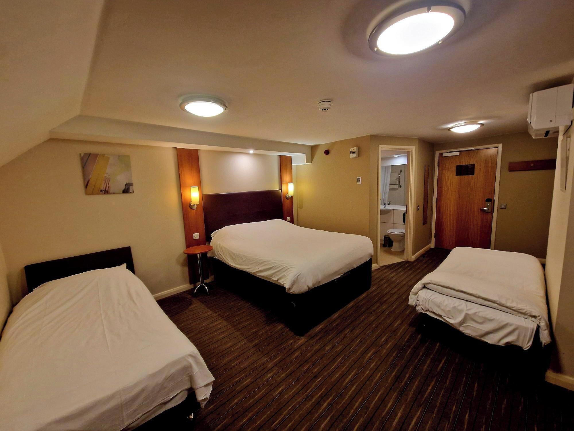 Habitación de hotel con dos camas, una mesita pequeña, una lámpara de pared, una luz en el techo y una puerta que da a un baño.