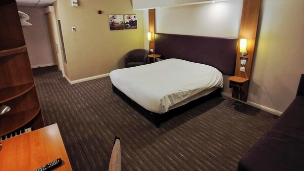 A hotel room with a bed, a desk, a chair, a small table, a nightstand, and a wall-mounted lamp. - Charter Hotel Bournemouth (Bournemouth)