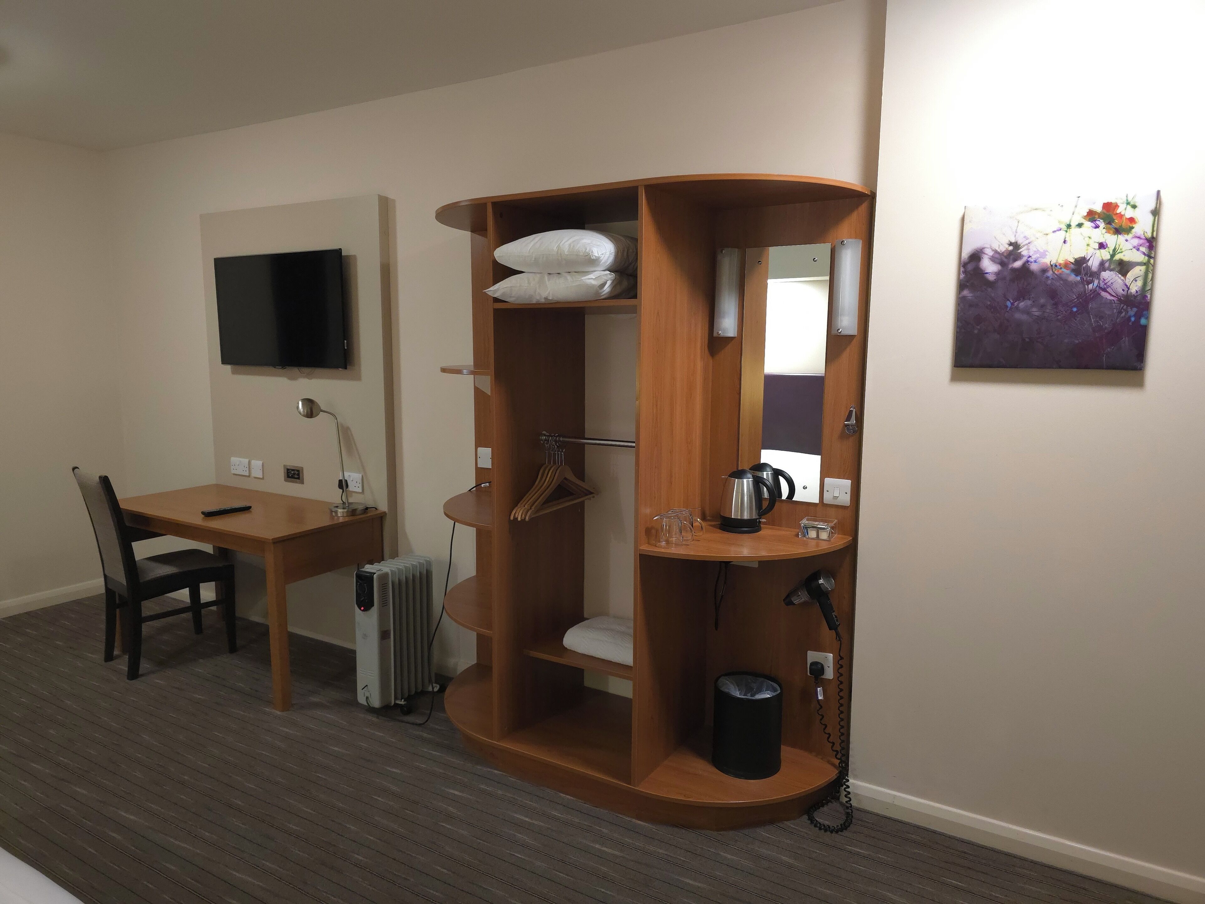 Habitación de hotel con escritorio de madera, una silla, un radiador, un televisor, un mueble de madera con espejo y un cuadro en la pared.