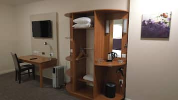 Habitación de hotel con escritorio de madera, una silla, un radiador, un televisor, un mueble de madera con espejo y un cuadro en la pared.