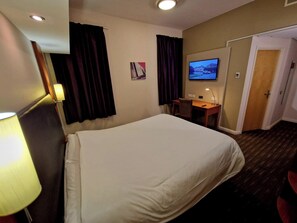 Quarto de hotel com uma cama, uma escrivaninha, uma televisão e uma porta.