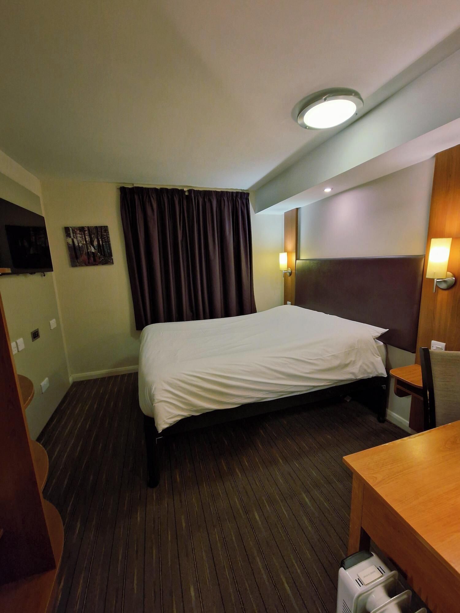 Ein Hotelzimmer mit Bett, Schreibtisch, Stuhl, Fernseher und Wandlampe.