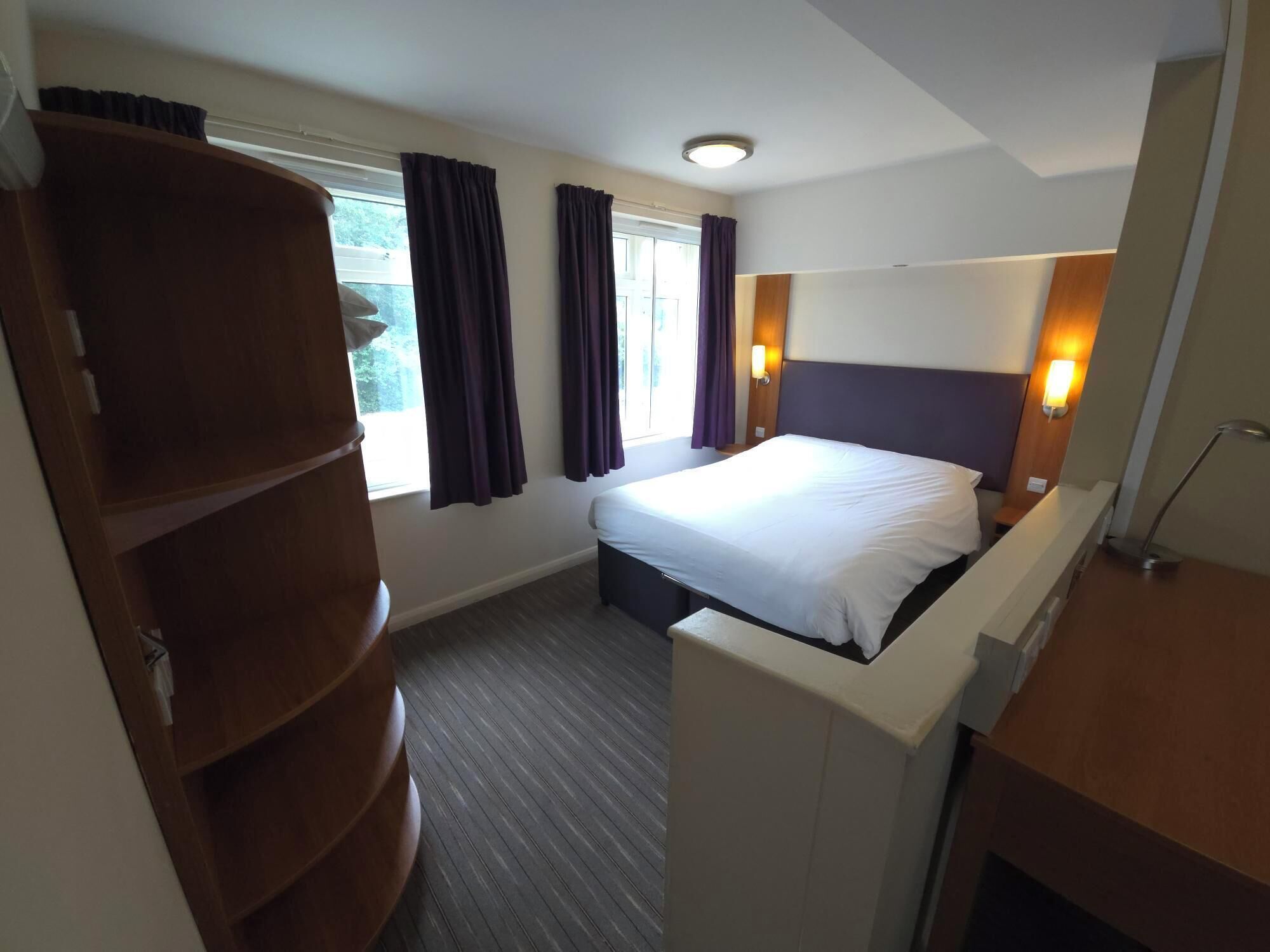 A hotel room with a bed, a wooden wardrobe, a desk, and a window with curtains.