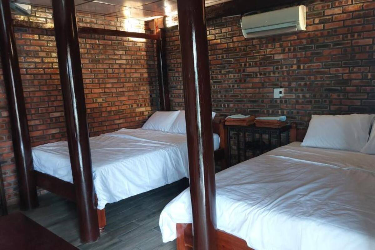 A room with two single beds and a larger bed, all with white linens. The room features exposed brick walls and a wooden ceiling.
