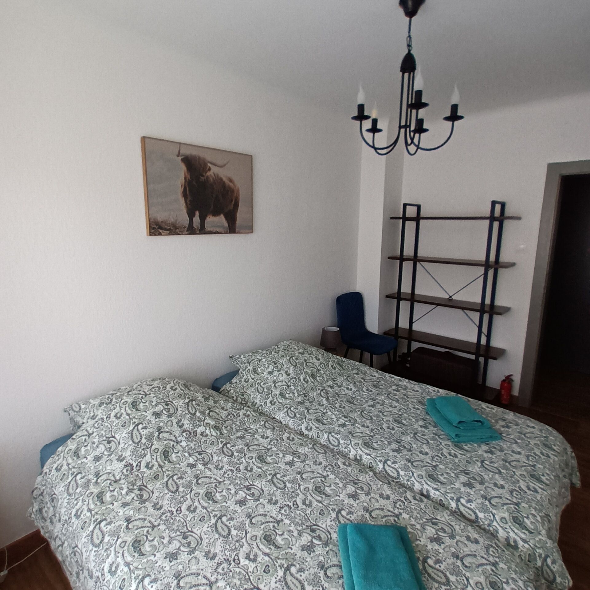 A bedroom with a bed, a chair, a chandelier, a painting, and a shelf.