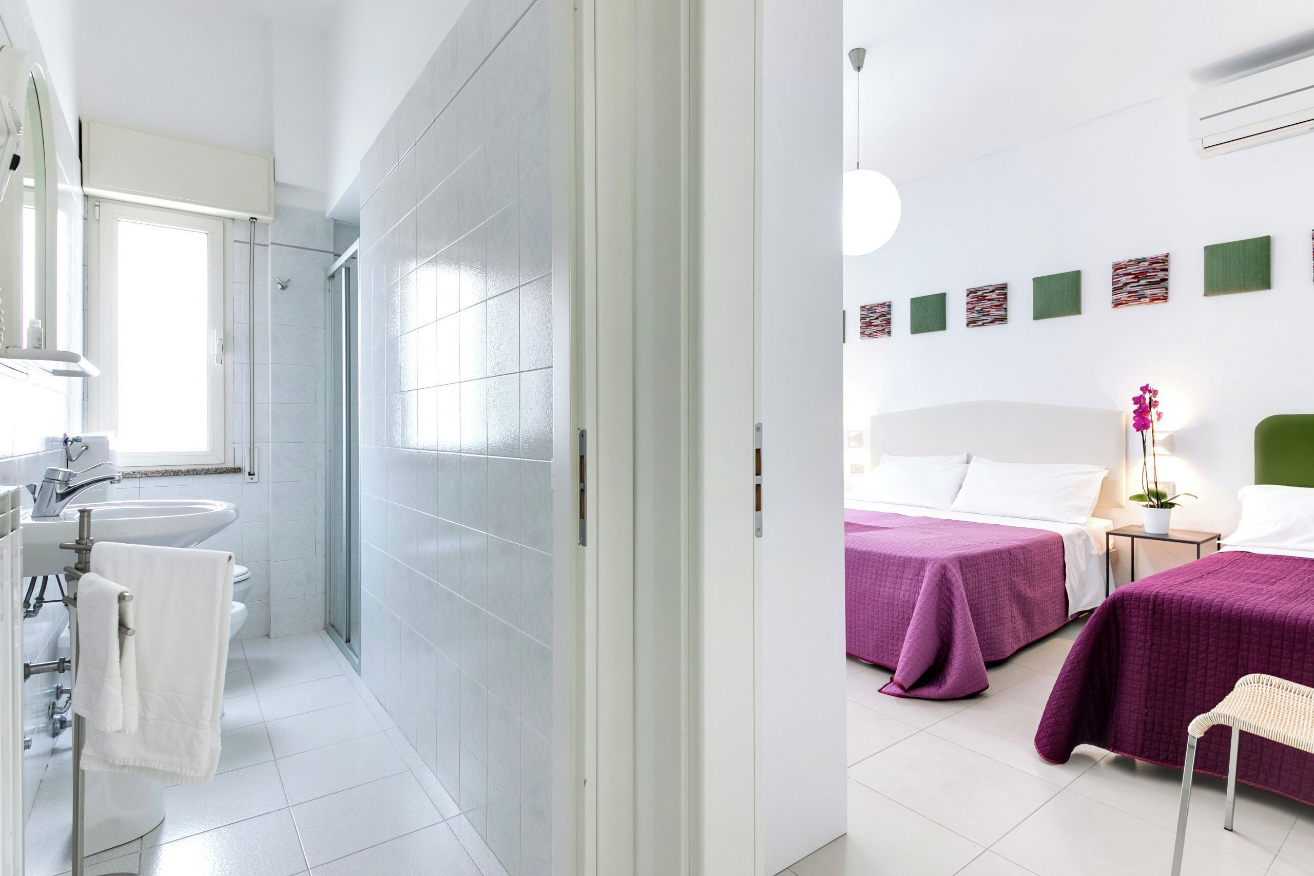 A modern bathroom with a shower, sink, and toilet. There is a bed with purple bedding and a nightstand with a lamp.