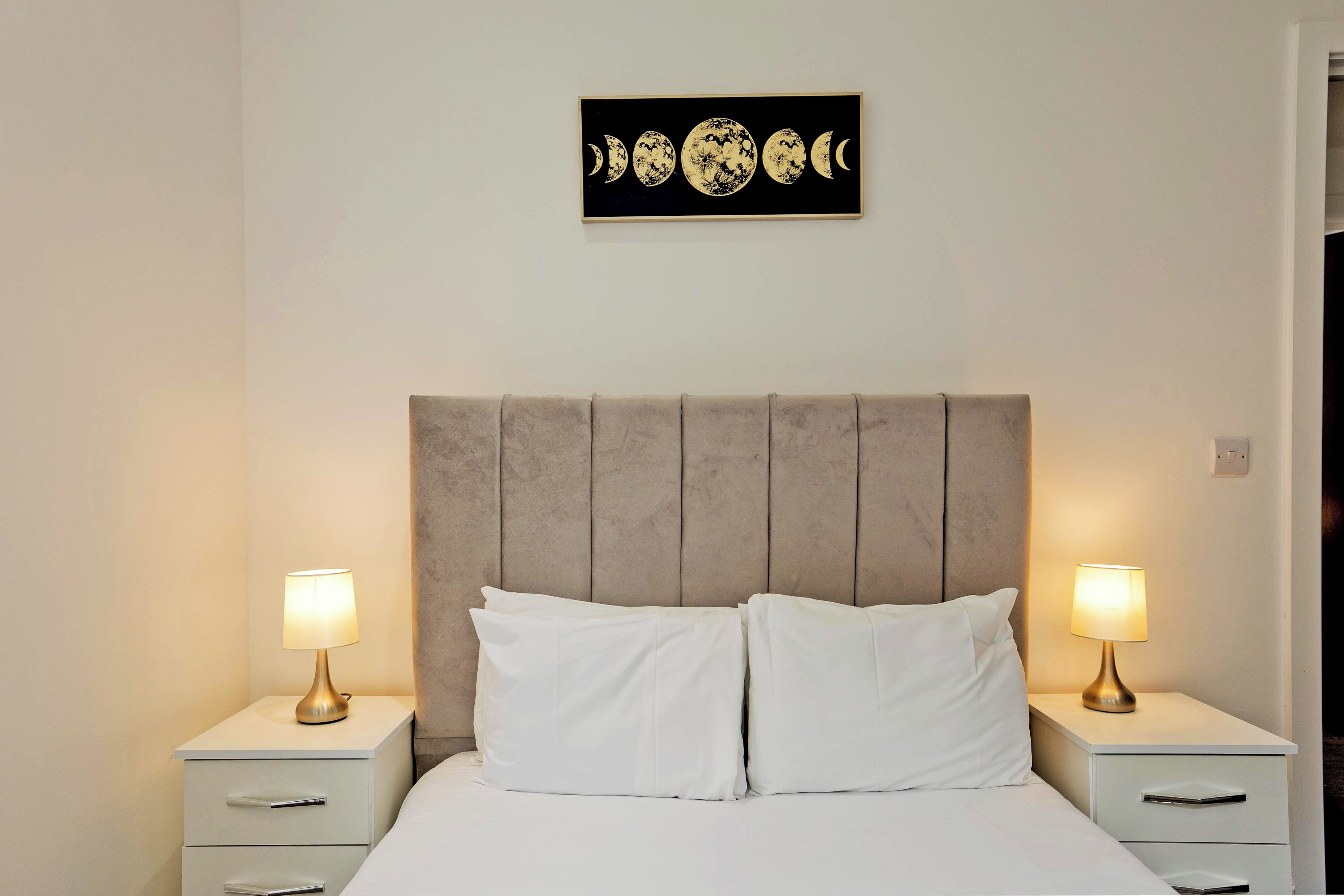 A bedroom with a bed, two bedside tables, a headboard, and a wall-mounted artwork depicting moon phases.