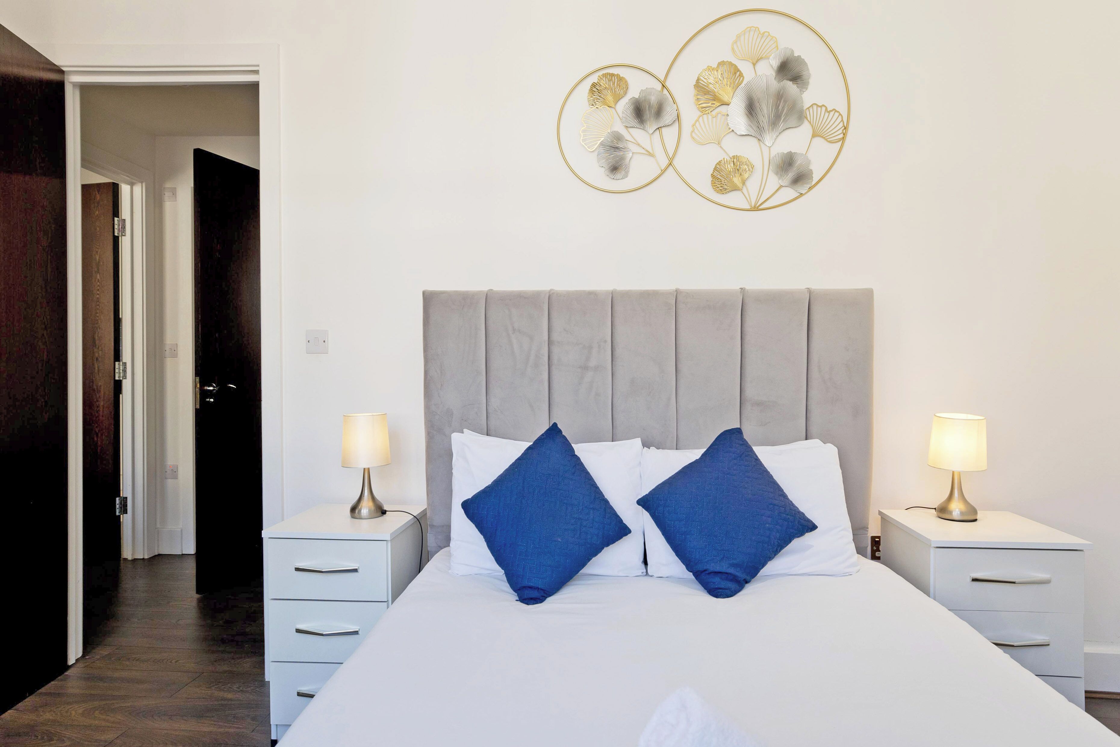 Habitación de hotel con una cama, dos mesitas de noche, un aparador, una obra de arte en la pared y una puerta que da a otra habitación.