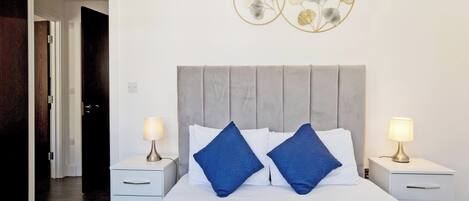 Habitación de hotel con una cama, dos mesitas de noche, un aparador, una obra de arte en la pared y una puerta que da a otra habitación.