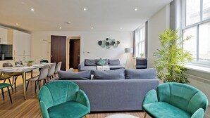 A modern living room with a grey sofa, teal armchairs, and a dining area with a table set for six. - Imperial Liverpool Street Apartments (London)