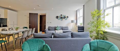 A modern living room with a grey sofa, teal armchairs, and a dining area with a table set for six.