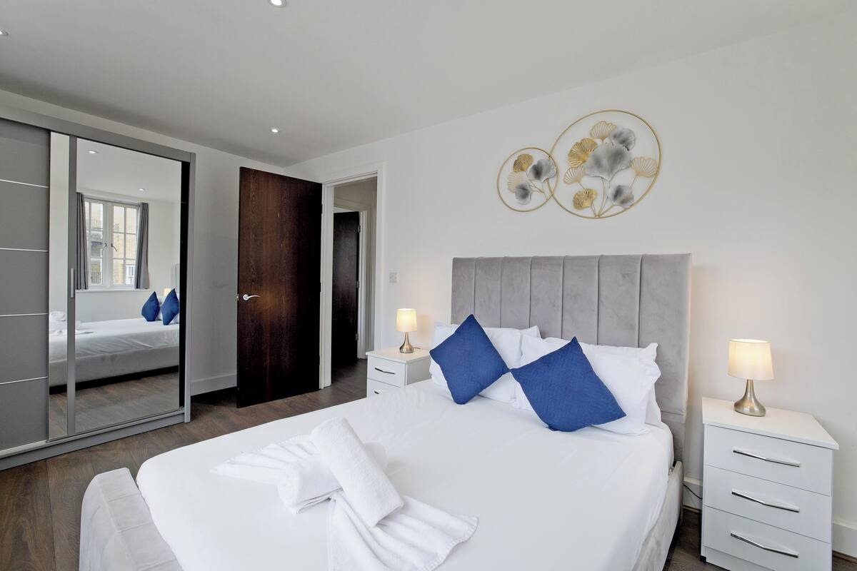 A modern hotel room with a large bed, a wardrobe, a bedside table with a lamp, and a mirror.