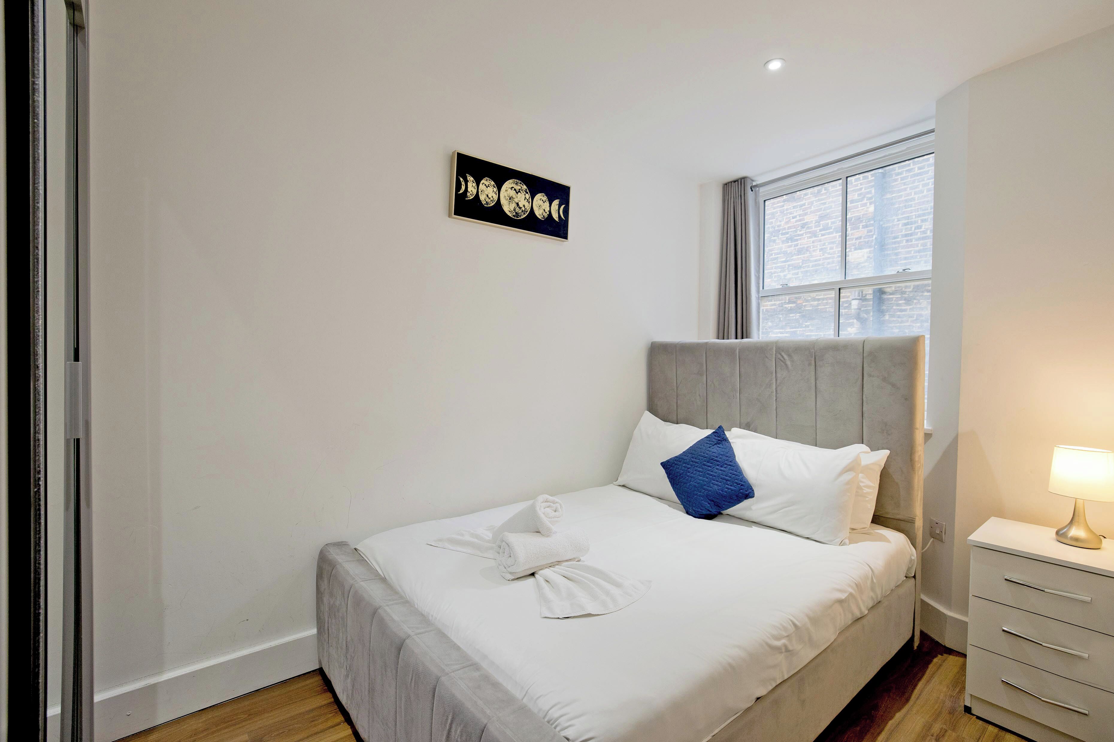 A neatly made bed with white linens and a blue pillow, a bedside table with a lamp, and a wall-mounted artwork depicting the moon phases.