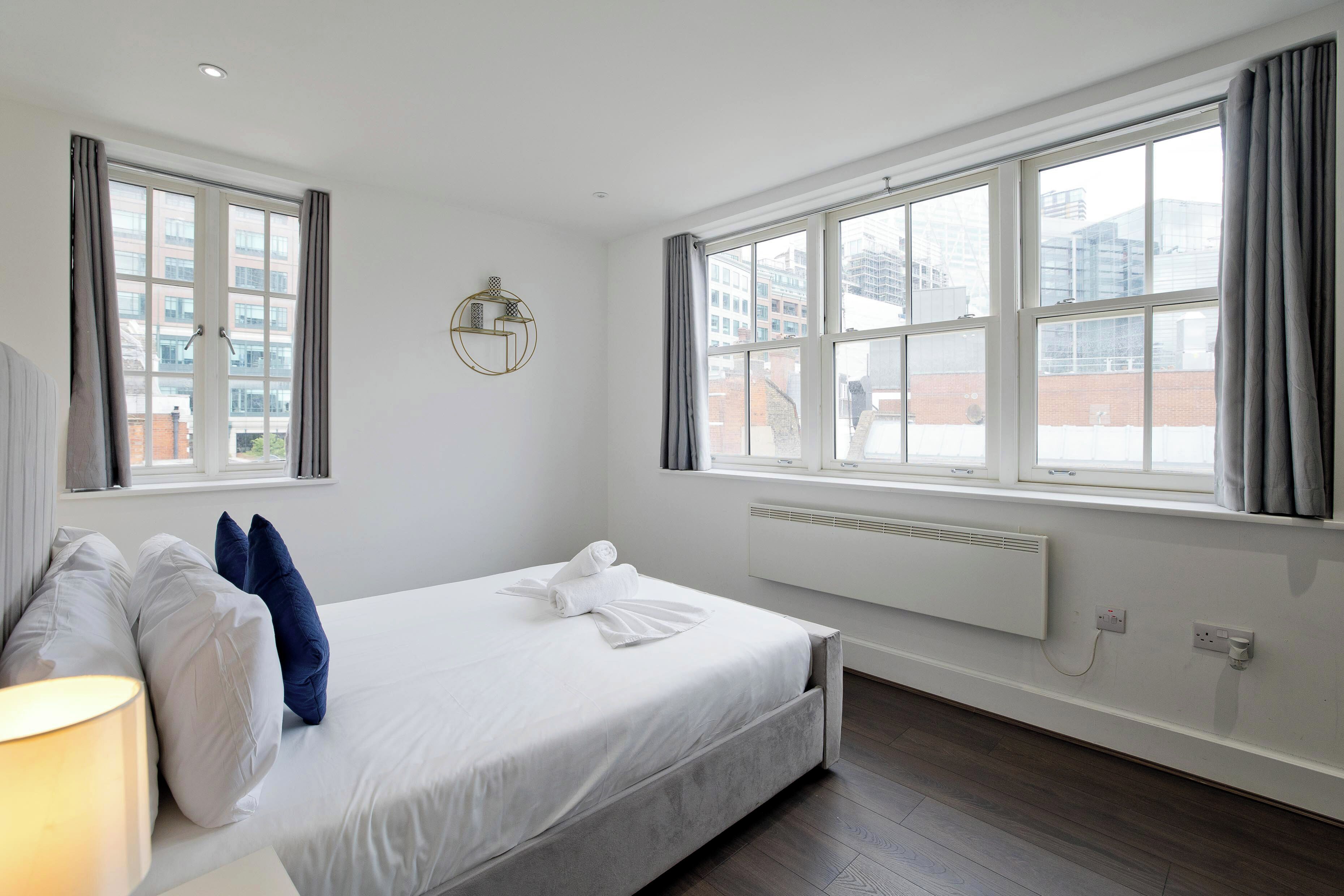 A hotel room with a large bed, two windows, a radiator, and a lamp.