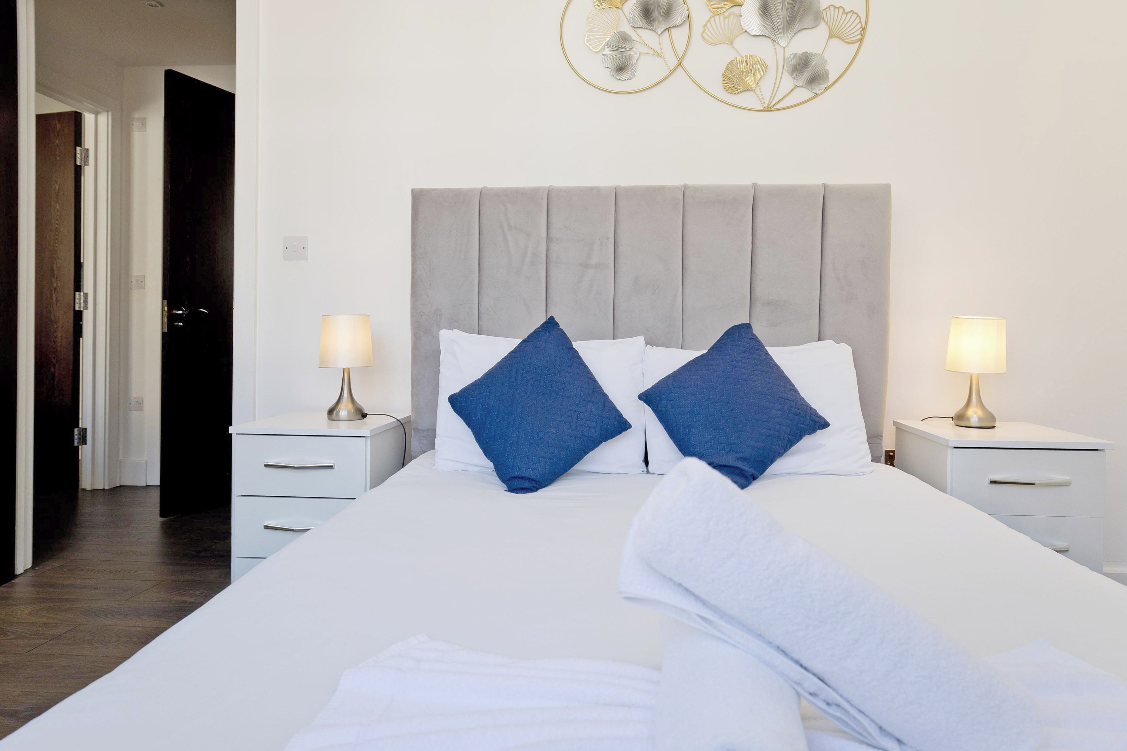 A neatly made bed with white and blue pillows, a grey upholstered headboard, and two bedside tables with lamps.