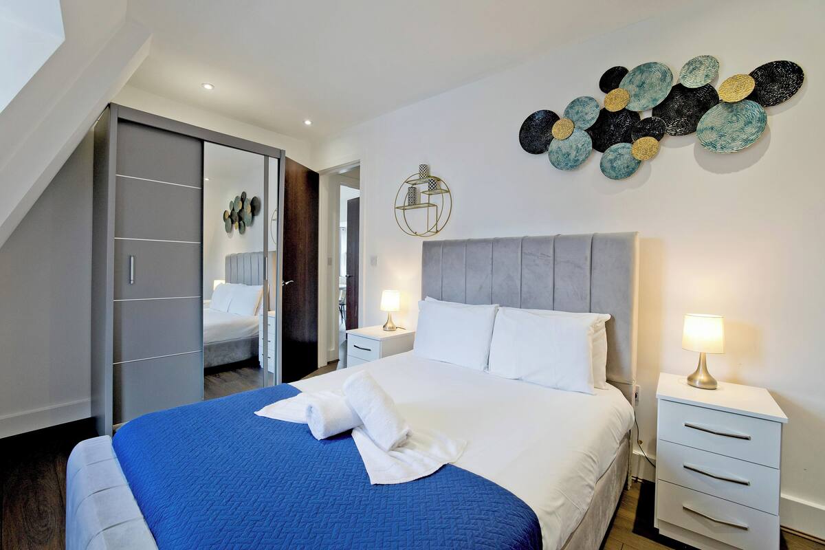 A modern hotel room with a large bed, a grey headboard, and a wall art piece. There is a bedside table with a lamp and a mirror.