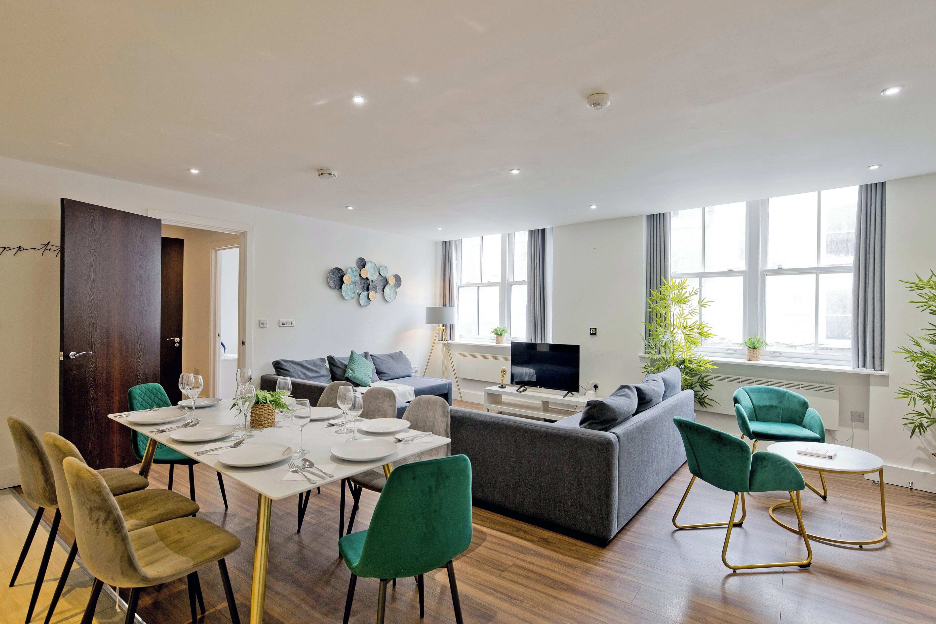 A modern living room with a dining area, grey sofas, green chairs, and a flat-screen TV.