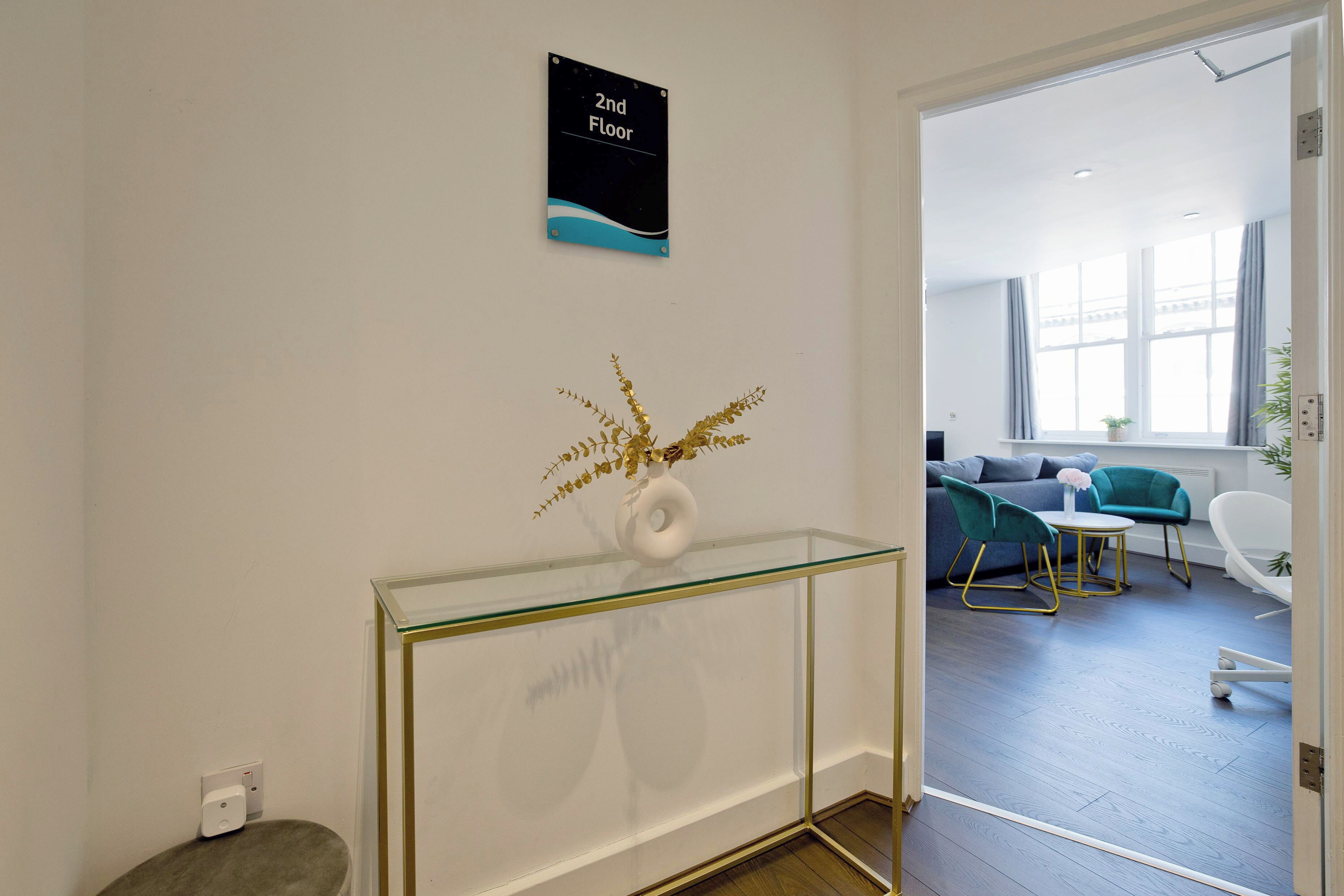 Un interior moderno con una consola de vidrio, un letrero que indica el segundo piso y un vistazo a una sala de estar con sofá y sillones.