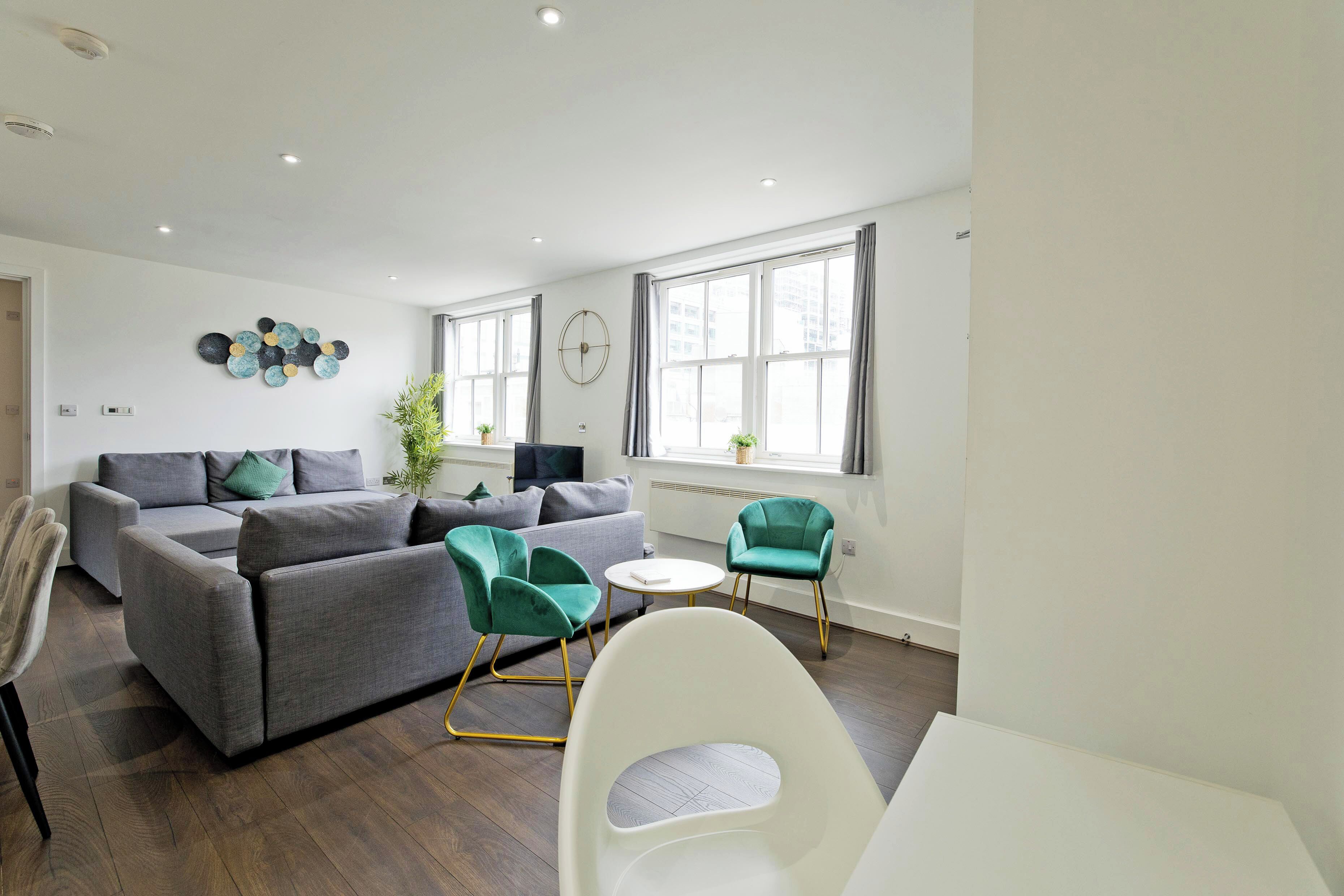 A modern living room with a grey sofa, teal chairs, a white table, and large windows.