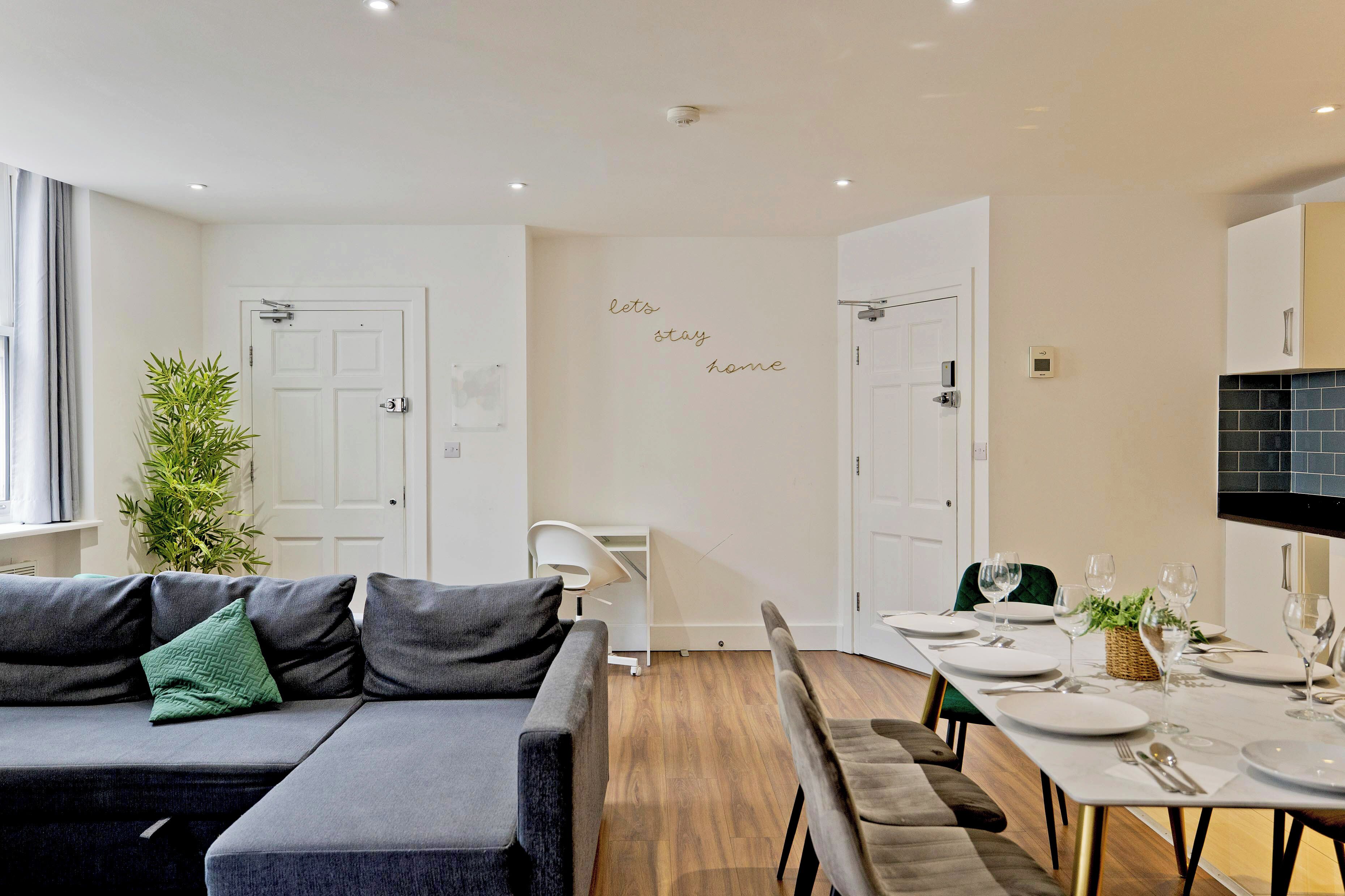 Een moderne woonkamer met een grijze bank, een eettafel voor vier personen en een keuken met een zwart betegelde achterwand.