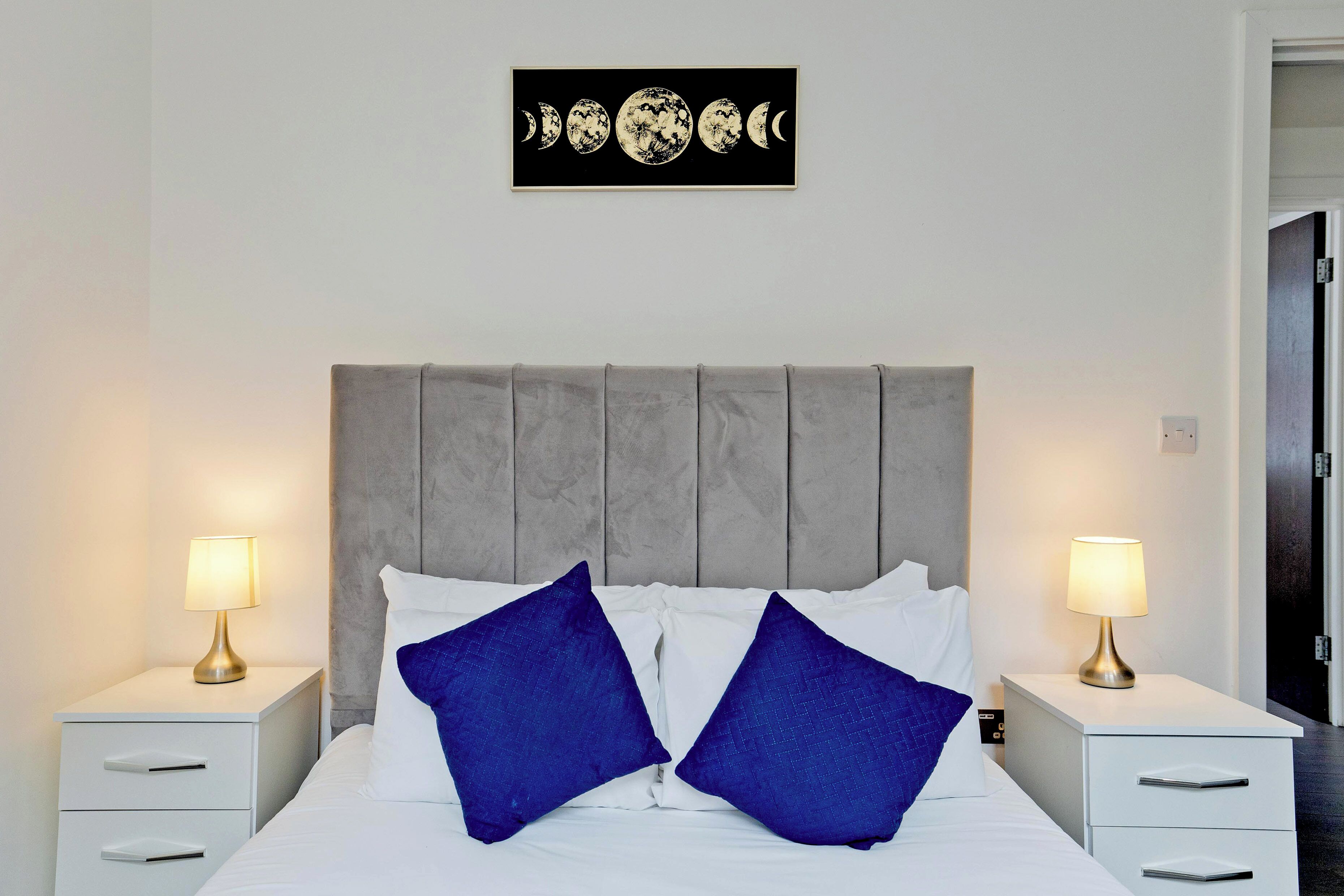 Un lit bien fait, avec deux oreillers bleus, une tête de lit grise et deux tables de chevet avec des lampes.