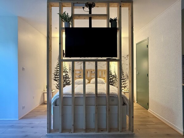 A bedroom with a bed, a TV mounted on a wooden partition, and Christmas trees in the background. - L'Échappée Ossaloise! (Eaux-Bonnes)