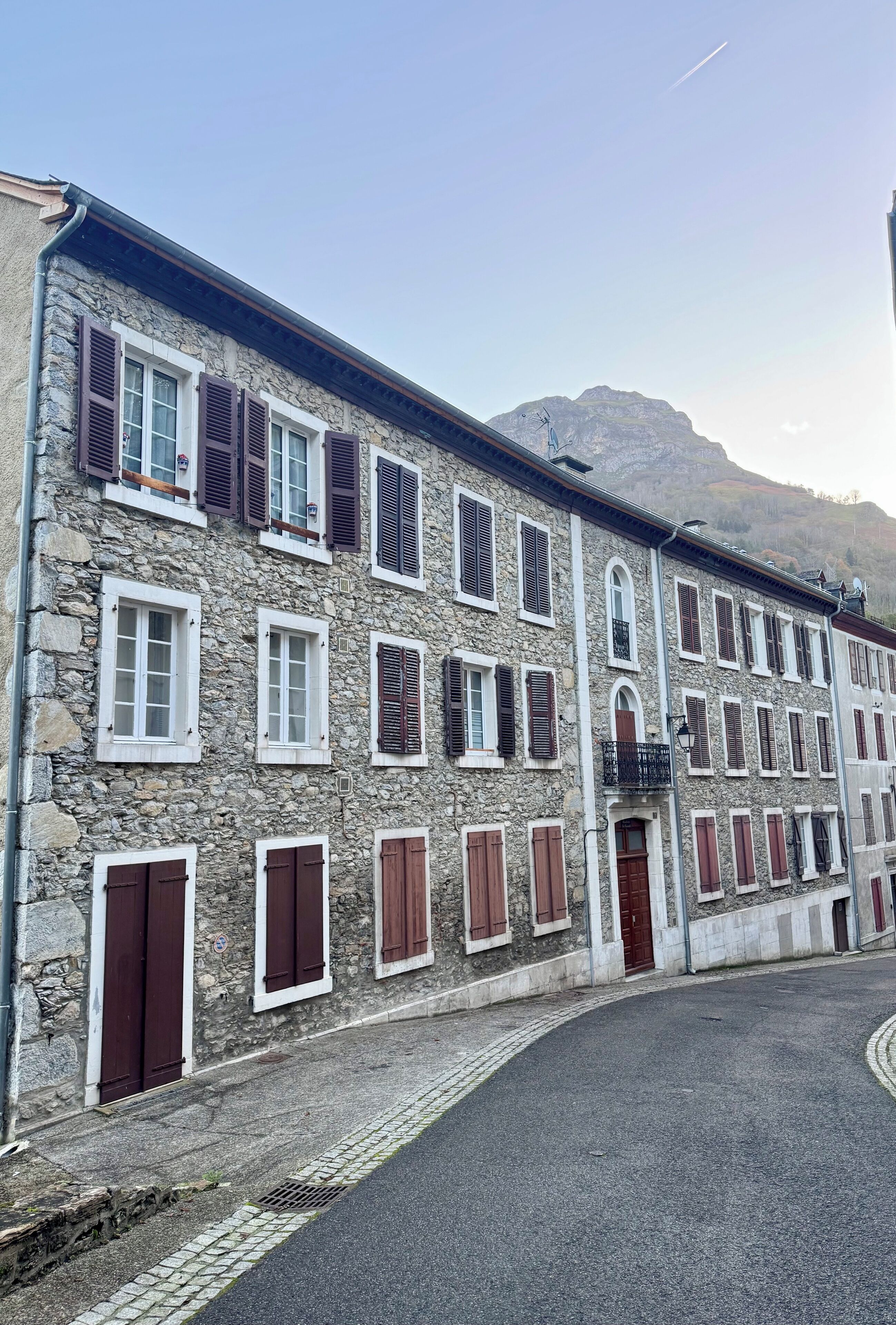 Une rangée de bâtiments en pierre, dotés de nombreuses fenêtres et portes, une rue pavée et une montagne en arrière-plan.