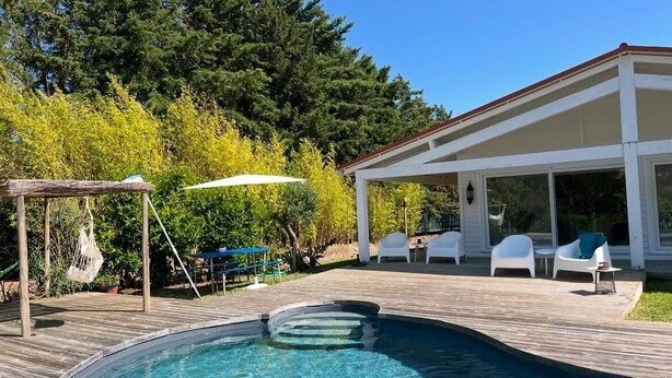 Une piscine dotée d’une terrasse en bois, entourée de chaises blanches et d’un espace abrité avec un mur en verre.