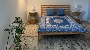 Une chambre à coucher avec un lit en bois, deux tables de chevet avec des lampes, une plante sur un support et une couvre-lit à motifs.
