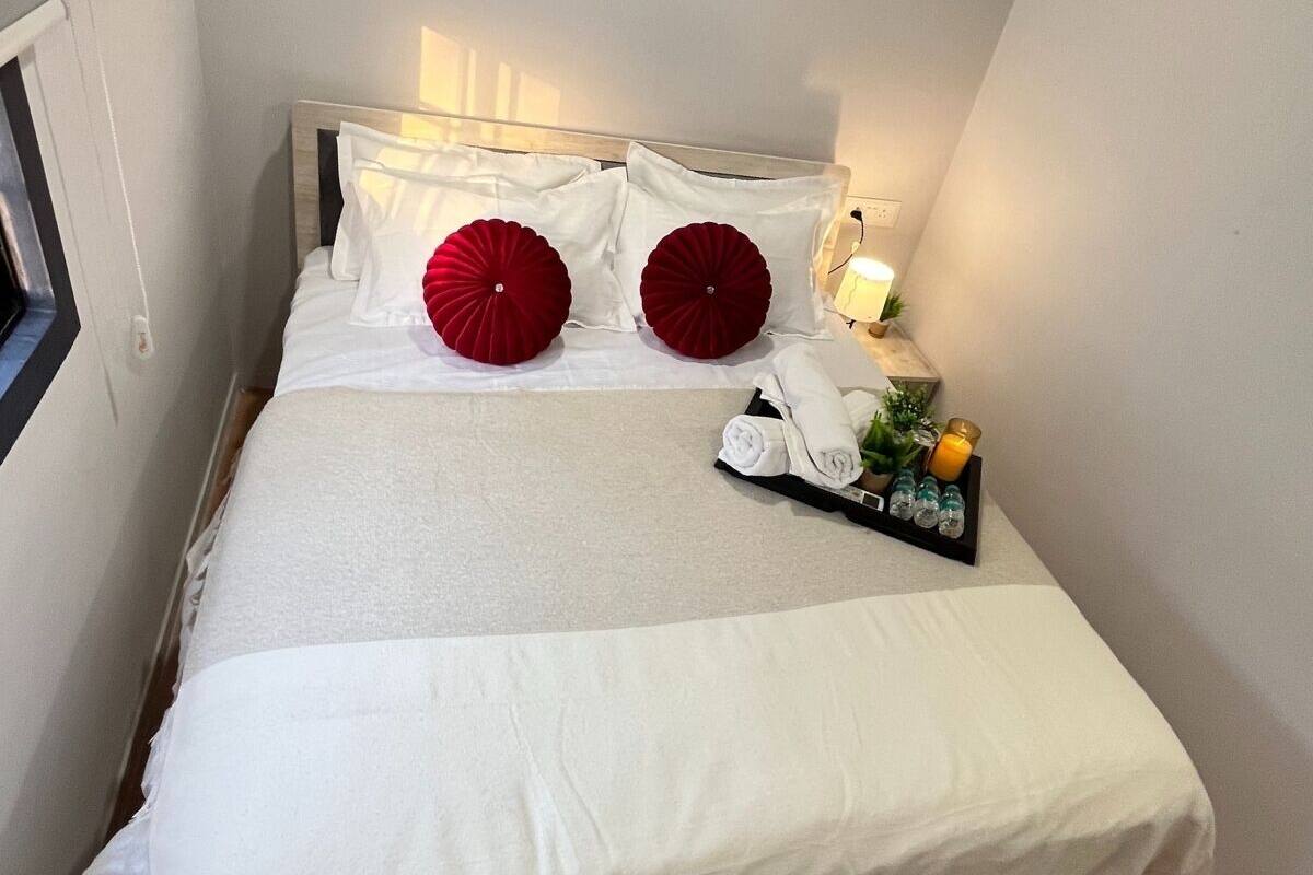 A neatly made bed with white linens and two red decorative pillows, a small bedside table with a lamp, and a tray with toiletries.