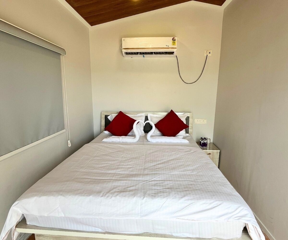 Una cama individual con una colcha blanca, dos almohadas rojas y un equipo de aire acondicionado blanco montado en la pared sobre la cama.
