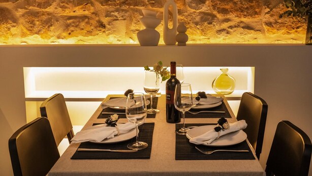 A dining table set with white plates, wine glasses, and a bottle of wine, with a stone wall and a vase in the background. - Montgo Loft Apartment (Dénia)