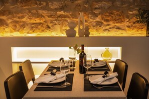 A dining table set with white plates, wine glasses, and a bottle of wine, with a stone wall and a vase in the background.