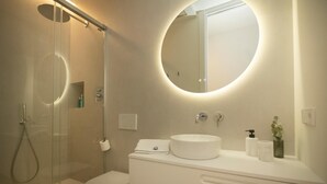 A modern bathroom with a glass-enclosed shower, a round mirror, and a white vanity with a sink. - Montgo Loft Apartment (Dénia)