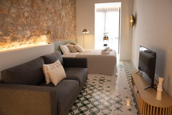 A modern hotel room with a stone wall, a sofa, a bed, a TV, and a patterned floor.