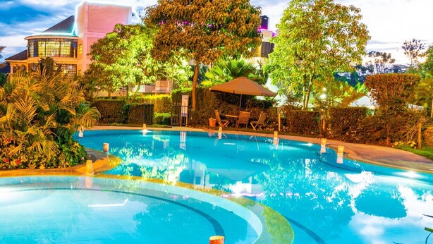 A swimming pool with surrounding lounge chairs and umbrellas, a landscaped garden, and a building with large windows in the background. - Royal Green Gardens (Nakuru)