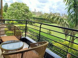 Un balcón con mobiliario de mimbre y una mesa de vidrio, con vistas a un jardín cuidado y edificios lejanos.