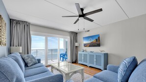 Un salon moderne avec un canapé bleu, une table basse en verre, un ventilateur de plafond et une vue sur l’extérieur grâce à de grandes fenêtres.