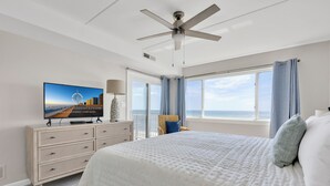 Une chambre avec un grand lit, un ventilateur de plafond, un téléviseur posé sur une commode et une vue sur l’océan par la fenêtre.