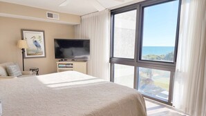 Une chambre d’hôtel avec un grand lit, un téléviseur à écran plat, une petite table, une lampe et une fenêtre donnant sur l’océan.