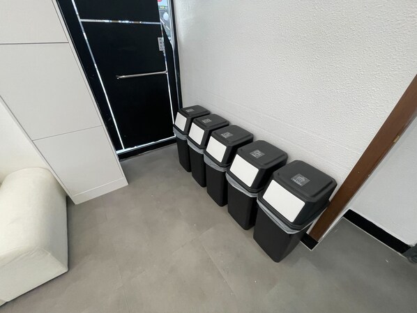 A corner of a hotel room with a white wall, a door, and four black trash bins with white labels. - SRR STAY (Gwangmyeong)