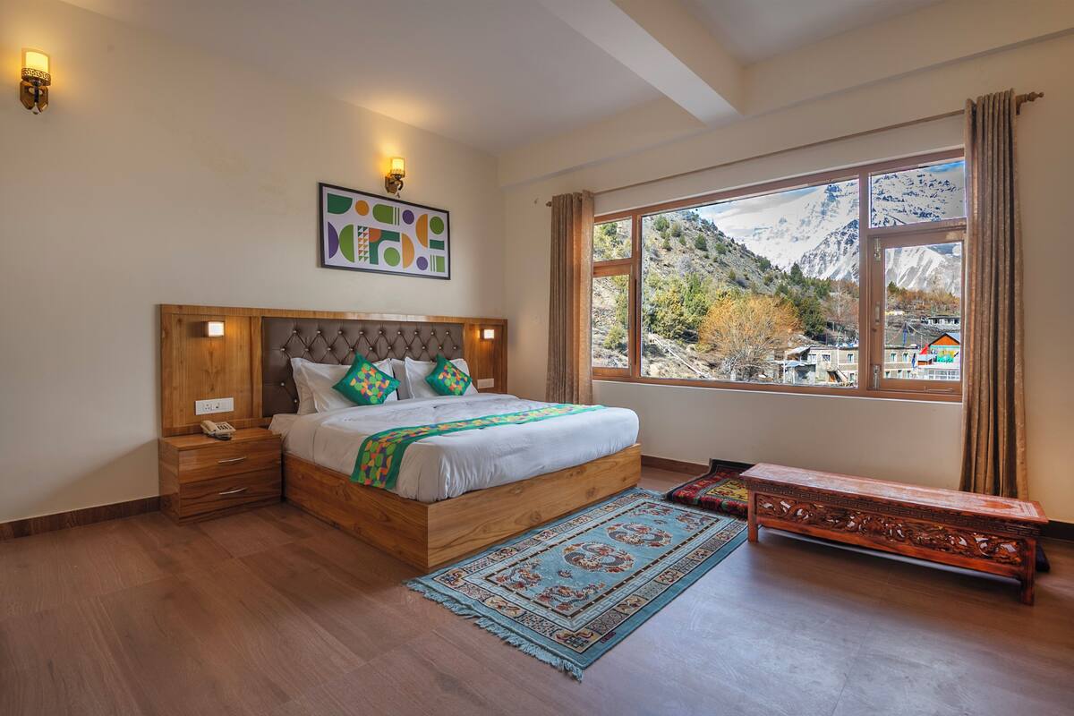 A bedroom with a wooden bed, bedside tables, a bench, a large window with a mountain view, and a wall-mounted artwork.