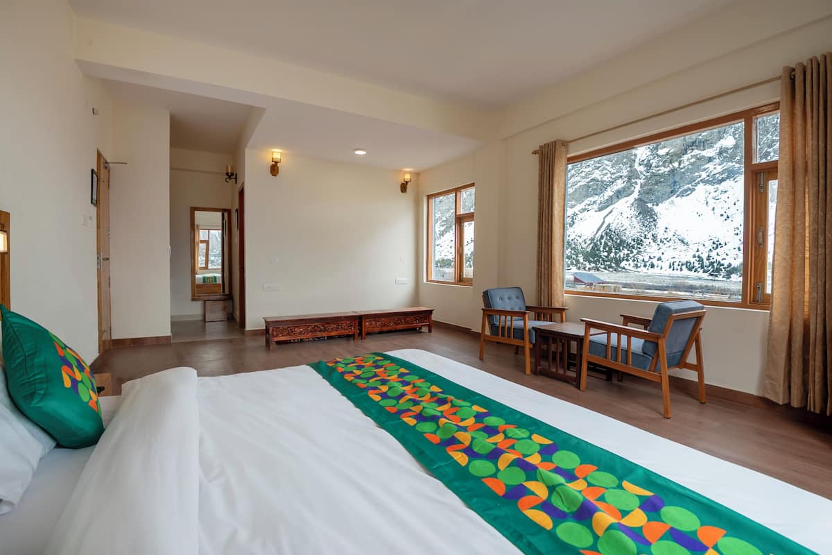 A hotel room with a bed, a desk, a chair, and a view of snow-capped mountains through large windows.