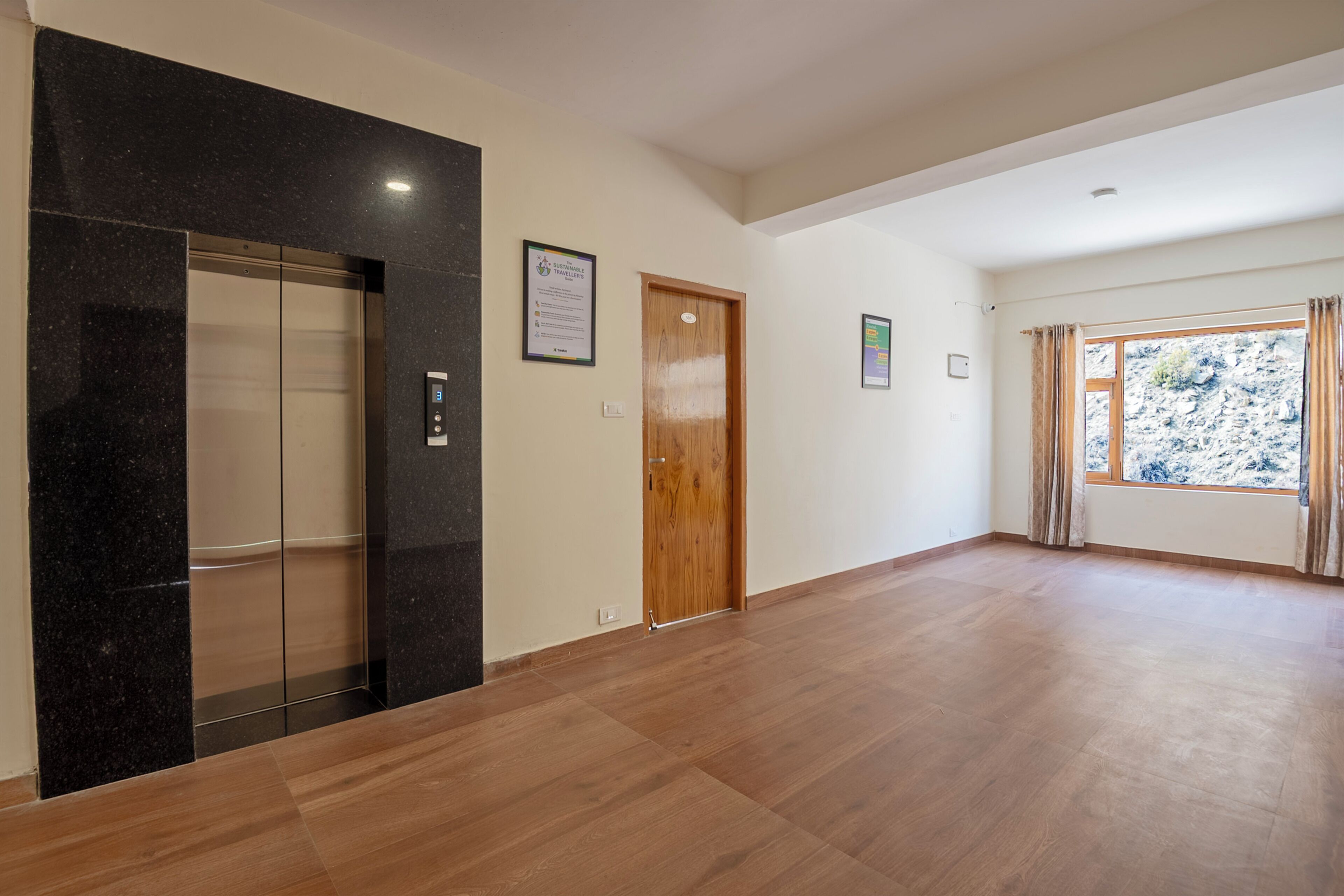 A spacious hallway with a wooden floor, a black elevator, a wooden door, and a window with curtains.