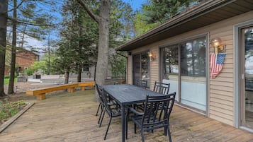 Une terrasse en bois avec une table et des chaises, entourée d’arbres et d’un bâtiment doté de grandes fenêtres et arborant un drapeau américain.