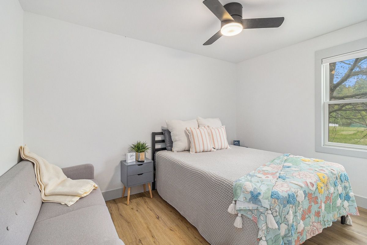 A bedroom with a bed, a grey sofa, a bedside table with a plant, a ceiling fan, and a window with a view of greenery.