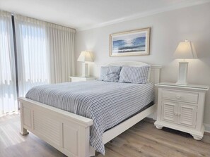 Un lit recouvert d’une couvre-lit à rayures, deux tables de chevet avec des lampes, un tableau encadré au mur et une fenêtre avec des rideaux légers.