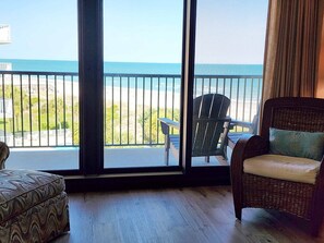 Una stanza con vista sulla spiaggia, una sedia in vimini, un poggiapiedi decorato e un'ampia finestra.