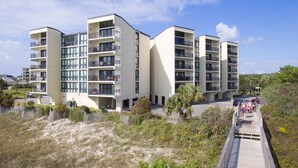 A modern multi-story hotel with balconies, located near a beach with a wooden boardwalk.
