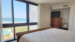 A hotel room with a large window offering a view of the beach and ocean, a bed with a white bedspread, a wooden dresser, and a television on top.